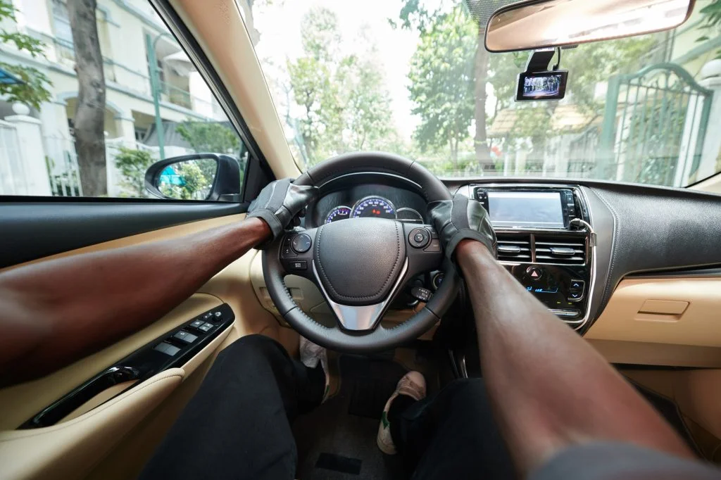 Driver's view of a person wearing gloves, gripping the steering wheel, while driving a car on a tree-lined street.