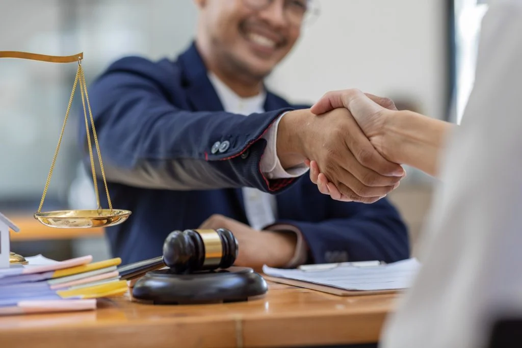 Two people shaking hands across a desk with a gavel and scales of justice, indicating a legal or professional agreement.