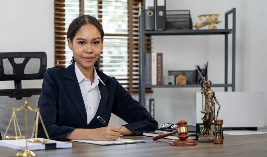 A woman in professional attire sits at a desk with legal materials, including the scales of justice, gavel, and Lady Justice statue, in an office setting.