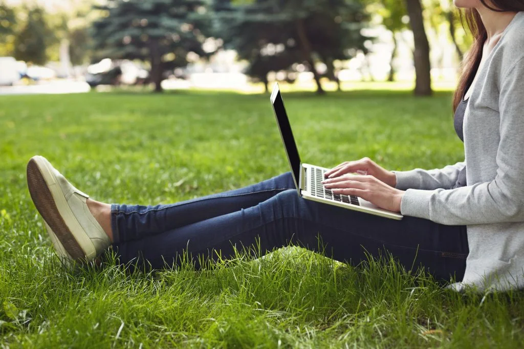 Person sitting on grass in a park, working on a laptop. Dressed in casual clothing with white shoes, jeans, and a light gray sweater. Trees and greenery are visible in the background.